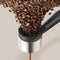 A stream of freshly ground beans cascades into the Philips Baristina Combo Espresso Machine with Milk Frother BAR303/60 (Black) on a light beige background. Brown coffee grounds are visible at the bottom, ready to create barista-quality espresso.