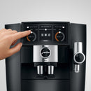 A close-up of a person's hand using the Jura J8 Twin Super Automatic Espresso Machine 15658 Diamond Black. The digital interface displays settings for "Broyeur gauche" and "Broyeur droite," featuring the P.A.G.2+ grinder. The person adjusts the left setting while the Jura J8 machine showcases two coffee spouts and a prominent brand logo.