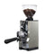 The Eureka Mignon Libra 65 All Purpose Grinder in chrome is an elegant coffee grinder that features Silent Technology and flat burrs. It comes with a transparent bean hopper filled with coffee beans, showcasing a digital display and a detachable portafilter holder, all presented against a plain white background.