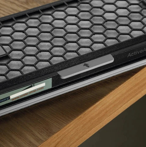 Close-up of a Miele Active AirClean filter with TimeStrip SF-AA 50 resting on a wooden surface. The filter, designed by the Miele brand, features an active charcoal honeycomb pattern and has a small directional arrow visible on its side.