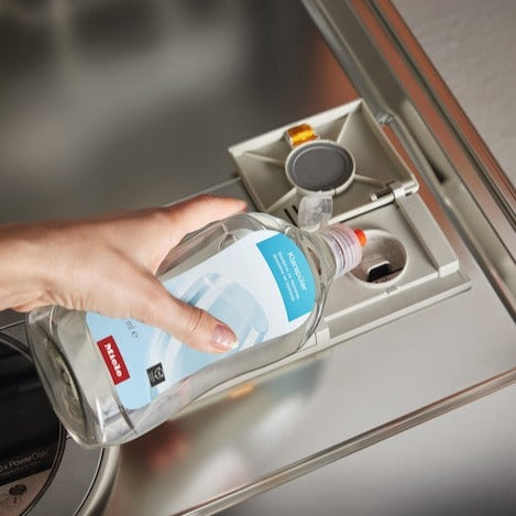 A hand is pouring Miele Dishwasher Rinse Agent 500 mL GS RA 503 L into the dispenser of a Miele dishwasher, prepping it for a QuickIntenseWash cycle. The detergent and rinse aid compartments are open and visible, ensuring sparkling shine with every wash.