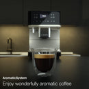 A double-walled glass cup filled with espresso sits on a countertop in front of a sleek, modern Miele CM6160 Milk Perfection Countertop Coffee Machine (Lotus White) with digital controls. The background shows a minimalistic kitchen setup. Text on the image reads, "AromaticSystem and OneTouch for Two. Enjoy wonderfully aromatic coffee.