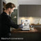 A man operates a Miele CM6160 Milk Perfection Countertop Coffee Machine (Lotus White) in a sleek, minimalist kitchen with beige and gray cabinetry. He presses a button on the machine, which features one-touch programming and a digital display. A glass of milk sits next to the coffee machine. The text reads "Maximum convenience.