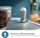 A Philips Barista Brew Semi Automatic Espresso Machine PSA3218/01 in sleek silver is placed on a metallic surface next to a dual bean container filled with assorted coffee varieties. The background is blurred, putting the focus on the espresso machine in the foreground. Text at the bottom reads, "Premium calibrated tamper for levelled coffee grounds.