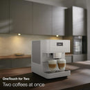 A sleek, modern Miele CM6160 Milk Perfection Countertop Coffee Machine (Lotus White) is on a kitchen counter, dispensing two cups of layered coffee simultaneously. The text on the image reads: "OneTouch for Two. Two coffees at once." The background shows a contemporary kitchen with soft lighting.