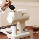 A person uses a black scoop to add coffee beans into a sleek, black Timemore Sculptor 064S All-Purpose Grinder on a wooden table. A small glass of espresso and an open book are nearby, creating a minimalist and cozy scene.