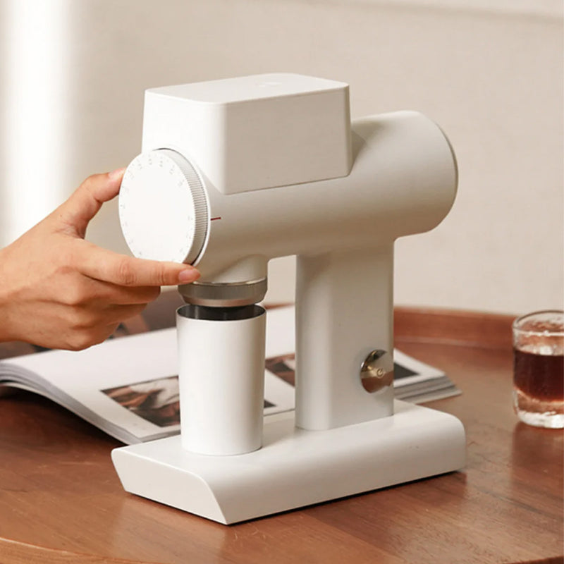 A person adjusts the settings on a modern, black Timemore Sculptor 064S All-Purpose Grinder resting on a wooden table. A small glass of espresso and an open book are in the background, showcasing its precise grinding capabilities.