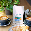 Equator Coffee Roasters North Star Coffee Bean (908g) sits between two black-cupped cappuccinos on a wooden table. In the foreground, a croissant joins the scene with a green potted plant and breakfast wrap, perfecting this medium-dark roast morning ritual.