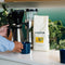 A person in a blue striped shirt pours coffee from a silver carafe into a blue mug on the counter. Next to them is a 2lb bag of Equator Coffee Roasters Sweet Justice Coffee Beans and green plants against a teal wall, enhancing the scene's vibrant harmony.