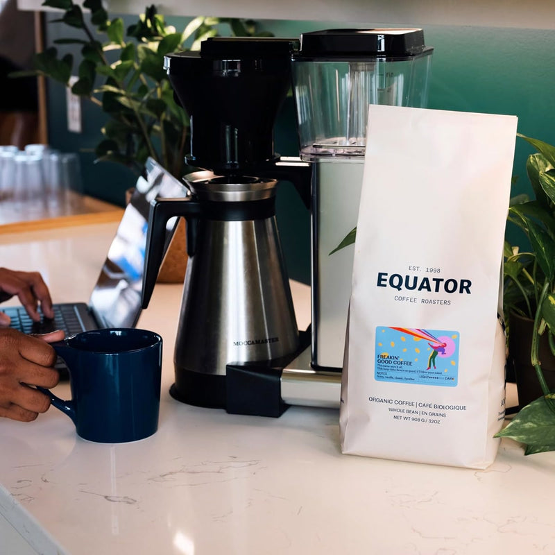 A person is using a laptop with their left hand while holding a blue mug filled with locally roasted, organic goodness in their right. Beside them on the white countertop are a coffee machine and Equator Coffee Roasters Freakin’ Good Coffee Bean (340g/12oz), poised for more brewing moments.