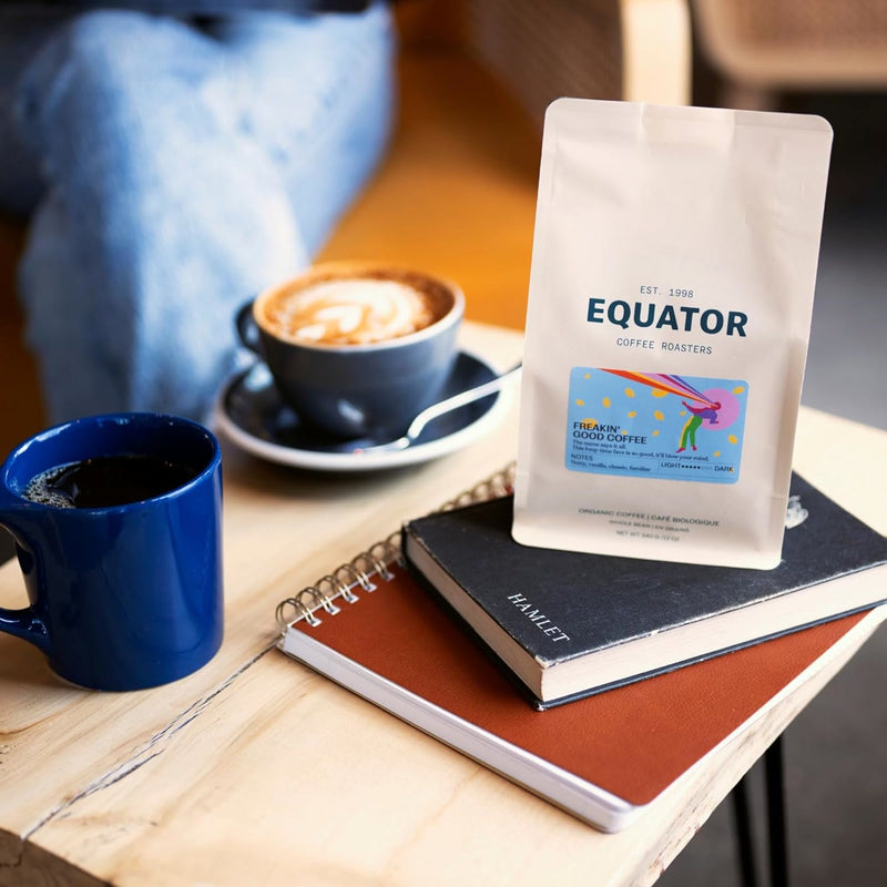 The table features a blue coffee cup, a latte in a black saucer, Equator Coffee Roasters Freakin’ Good Coffee Bean (908g / 2lb), a spiral notebook, and "Hamlet." A blurred arm on a chair can be seen in the background.