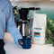 In a kitchen, someone in a blue striped shirt holds a blue mug. A bag of Equator Coffee Roasters North Star Coffee Beans (908g / 2lb) rests on the counter with a coffee maker, exuding its medium-dark roast aroma. A green plant adds nature to the scene.