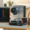 A person holding a Nespresso cup stands beside a Nespresso Momento 100 Commercial Coffee Machine on a counter. The machine features touchless technology, a digital display, and a single spout for dispensing coffee. In the background is a Nespresso capsule dispenser filled with colorful coffee pods.