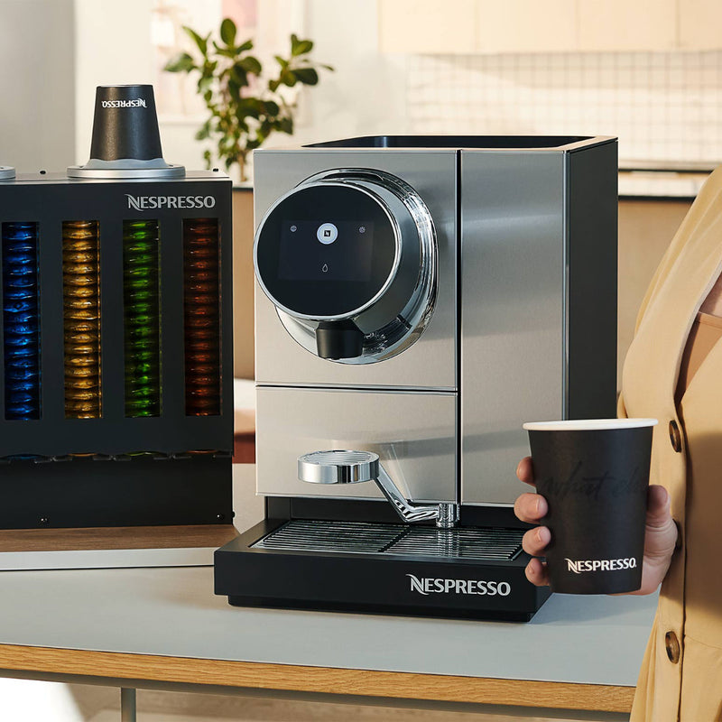 A person holding a Nespresso cup stands beside a Nespresso Momento 100 Commercial Coffee Machine on a counter. The machine features touchless technology, a digital display, and a single spout for dispensing coffee. In the background is a Nespresso capsule dispenser filled with colorful coffee pods.