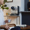 A person sits in a chair holding a Nespresso cup in their right hand. The Nespresso Momento 100 Commercial Coffee Machine from Nespresso, equipped with capsule recognition, sits on the table beside them, next to a plant and a jar of colorful items. Another person wearing black stands near the countertop in the background.