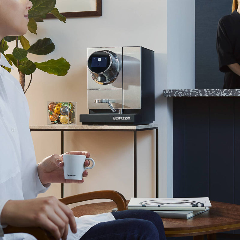 A person sits in a chair holding a Nespresso cup in their right hand. The Nespresso Momento 100 Commercial Coffee Machine from Nespresso, equipped with capsule recognition, sits on the table beside them, next to a plant and a jar of colorful items. Another person wearing black stands near the countertop in the background.