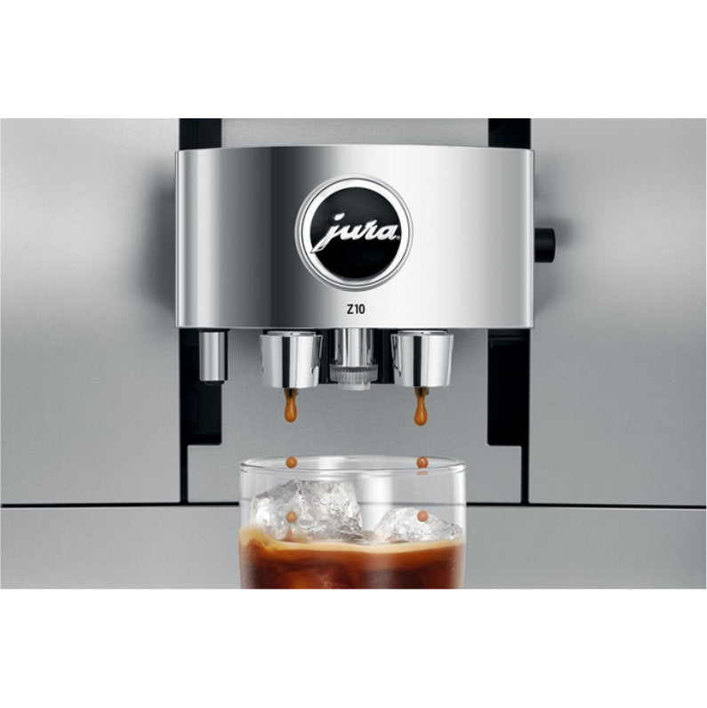 A close-up of the Jura Z10 Aluminum White Super Automatic Hot Coffee & Espresso, Cold Brew, & Specialty Beverage Machine - DEMO (4082), a marvel of Swiss engineering, dispensing coffee into a glass filled with ice. The aluminum white machine showcases its precision as the coffee is shown mid-pour, droplets falling delicately into the glass—perfect for cold brew specialties.