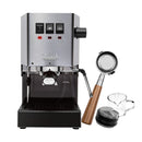 The Gaggia Classic E24 Espresso Machine RI9380/46, known for its sleek stainless steel design, includes a bottomless walnut portafilter. It comes bundled with a small measuring glass and distributor. Featuring the iconic "Gaggia" branding on the front, this model has an intuitive control panel with three buttons.