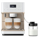 The Miele CM6360 Super Automatic Countertop Coffee & Espresso Machine in Lotus White features two cappuccino glasses filled with layered coffee and froth. A milk container connects to the machine via a tube, indicating automated milk frothing. The digital display is visible at the top.