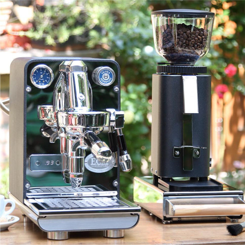 An ECM Puristika Semi Automatic Espresso Machine in anthracite, equipped with an E61 group head, and an ECM C-Manuale 54 Burr Grinder in the same shade rest on a wooden table outdoors. This semi-automatic espresso machine includes a pressure gauge and steam wand, while the grinder boasts a hopper filled with coffee beans. A small cup is partially visible on the left next to this elegant setup.