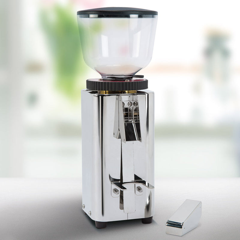 Introducing the ECM C-Manuale 54 Burr Grinder by ECM, a sleek stainless steel device with a transparent top, ideal for freshly ground coffee beans. This on-demand grinder features a boxy design accompanied by a small detached rectangular piece. It is beautifully showcased against a softly blurred background.