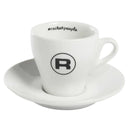 A set of six white ceramic espresso cups with handles, part of the Rocket Espresso Cups Hashtag Series - Set of 6 RA99907206 (White) from Rocket, sits on matching saucers. Each cup features a black circle with a bold letter "R" in the center and the hashtag "