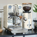 A sleek espresso machine with a metallic finish and wood accents, featuring a pressure gauge, sits on a black countertop. Beside it is the ECM C-Manuale 54 Burr Grinder in cream, filled with coffee beans and offering graduated grind adjustments. A potted plant and glass of water add to the ambiance in the background.