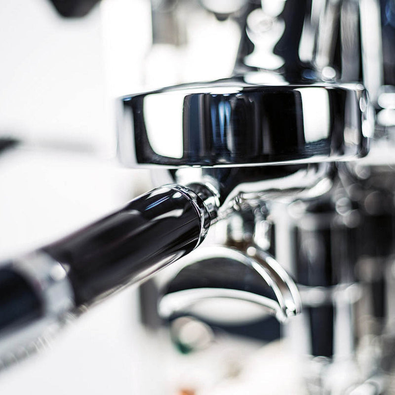 Close-up shot of the ECM Mechanika VI Slim Espresso Machine - Demo, Unused's portafilter and group head, with a blurred background, showcasing the stainless steel construction and black handle components. The focus is on the polished metallic parts, highlighting their reflective surfaces ideal for coffee brewing and milk frothing.