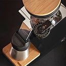 A top-down view of the Eureka Oro Mignon Single Dose Pro Grinder in black showcases its elegant metal and wood design. Coffee beans sit inside, poised for the flat burrs to transform them. A numbered dial with a small red indicator offers precise grind size adjustment. The "Eureka" logo adorns both the base and the collection cup.