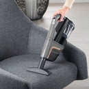 A person uses a Miele vacuum cleaner equipped with the Miele Wide Upholstery Nozzle Vacuum Cleaner Attachment SPD 20 to clean the seat of a gray upholstered chair. The vacuum features a sleek design and a transparent dust compartment, with a decorative vase visible in the background on a light wood floor.