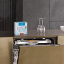 A dishwasher with an open door is built into a modern, minimalist kitchen. Two wine glasses are placed upside down on the countertop beside it. A box of Miele UltraTabs All in 1 Dishwasher Cleaning Tablets (Box of 60) sits on the counter above the dishwasher, ready to deliver powerful cleaning with QuickIntenseWash.