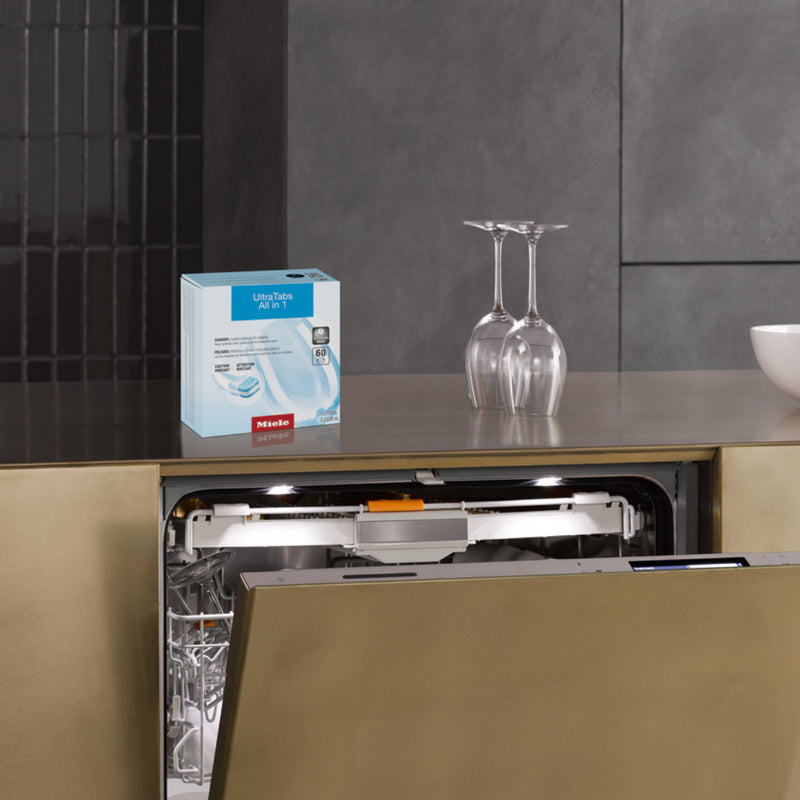 A dishwasher with an open door is built into a modern, minimalist kitchen. Two wine glasses are placed upside down on the countertop beside it. A box of Miele UltraTabs All in 1 Dishwasher Cleaning Tablets (Box of 60) sits on the counter above the dishwasher, ready to deliver powerful cleaning with QuickIntenseWash.