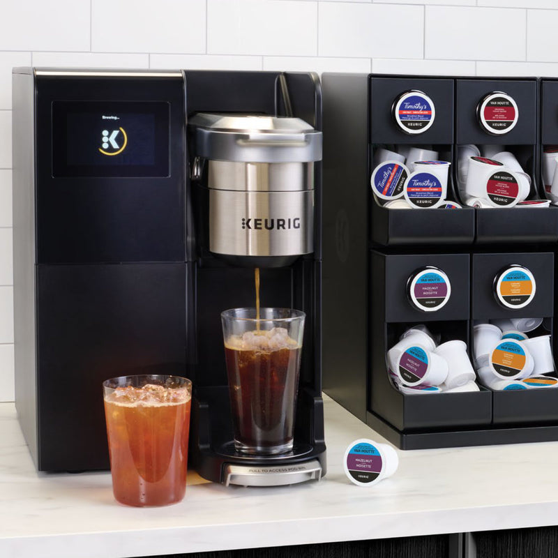 A Keurig K3550 K-Cup® Commercial Brewing System brews refreshing iced coffee into a glass filled with ice while another glass of chilled coffee sits on the counter. In the background, a storage rack displays an array of Keurig coffee pods.