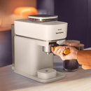 A person is using a white Philips Baristina Combo Espresso Machine with Milk Frother BAR303/00 placed on a counter. They are holding the portafilter handle, preparing barista-quality espresso with freshly ground beans. In the background, there are a jar and some other items on the counter.