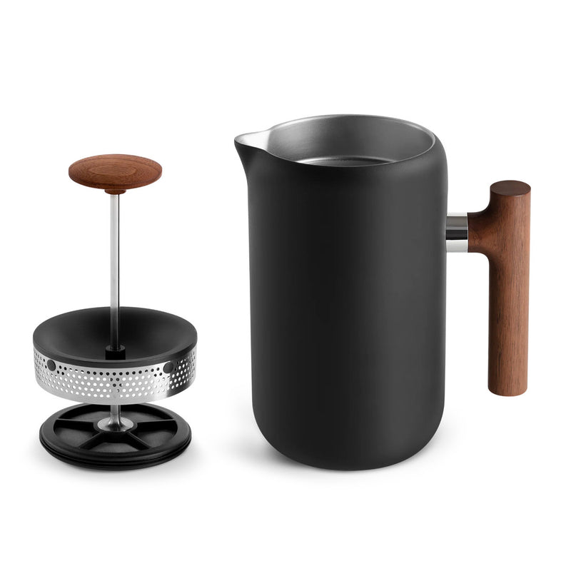 The Fellow Clara French Press (Matte Black + Walnut) by Fellow is showcased disassembled, highlighting its barista-level functionality. The sleek cylindrical pot sits next to the stainless steel plunger assembly, which features a mesh filter and a wooden knob, all embodying a minimal design.