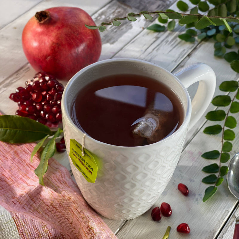 A white mug filled with Bigelow Green Tea with Pomegranate and a tea bag from the box sits on a wooden surface. Surrounding it are a pomegranate, its seeds, green leaves, and a pink cloth. The scene is beautifully illuminated by natural light.