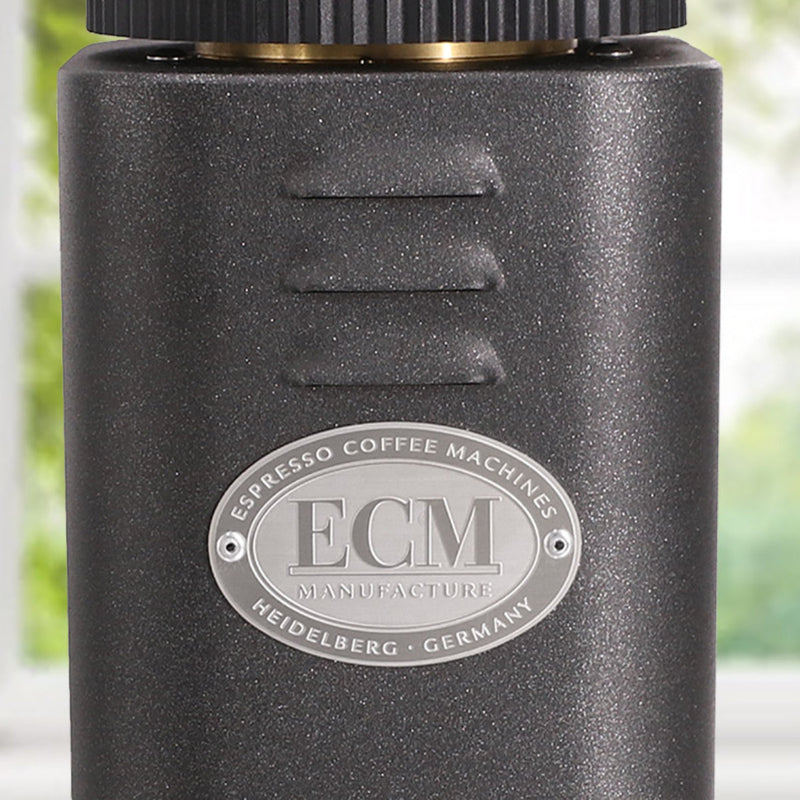 A close-up of a dark metallic ECM espresso coffee machine prominently features the brand's logo. The oval emblem, highlighting "Espresso Coffee Machines" and "Heidelberg - Germany," showcases a seamless design ideal for pairing with the ECM C-Manuale 54 Burr Grinder in Anthracite. Soft lighting accentuates the machine's texture.