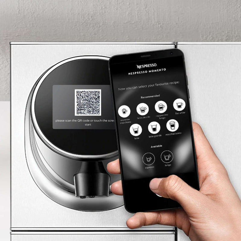 A person is holding a smartphone displaying the Nespresso app in front of a Nespresso Momento 100 Commercial Coffee Machine. The machine features a circular interface with capsule recognition and touchless technology. The phone screen shows multiple coffee options, including espresso and lungo.