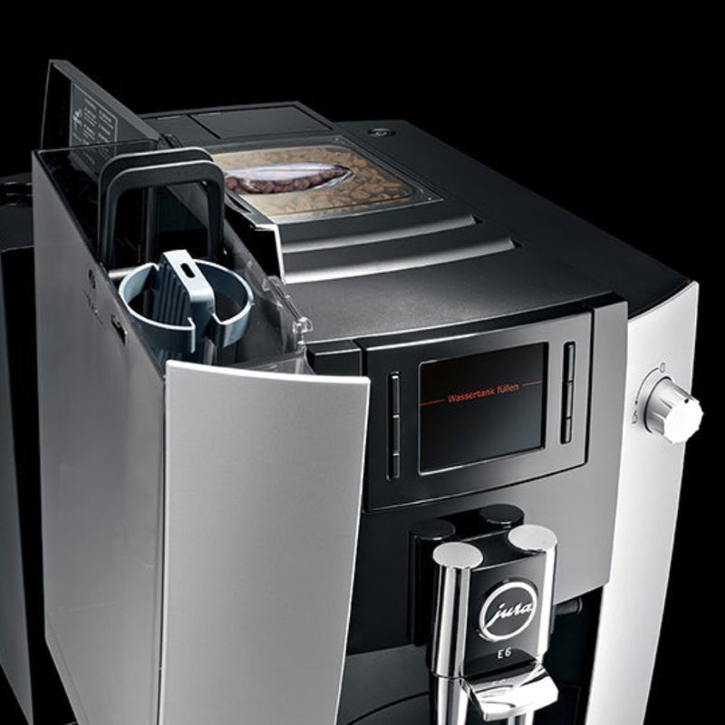 The Jura E6 Super Automatic Espresso Machine 15070 (Platinum) by Jura flaunts a modern, sleek silver and black design. It comes equipped with a digital display screen and an AromaG3 Grinder to enhance the coffee's flavor. There's a coffee bean container positioned on top, with space for your cup below the dispenser. The open side compartment reveals a spacious water tank, adding to its functional style.