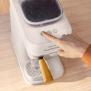 A person presses a button on a Philips Baristina Combo Espresso Machine with Milk Frother BAR303/00 (White), featuring a yellow portafilter and a clear bean hopper on top. The scene suggests the initiation of the coffee brewing process with fresh ground beans against a light wood surface background.