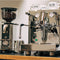 An ECM Mechanika Max Heat Exchanger (HX) Espresso Machine with PID in stainless steel, featuring sleek knobs and dials and a precise brew temperature control system, sits on a counter. Behind it stands the ECM S-Automatik 64 Burr Grinder, also in stainless steel, filled with beans. Two white cups rest on top of the espresso machine, alongside a milk frothing pitcher from the Barista Tools Bundle.