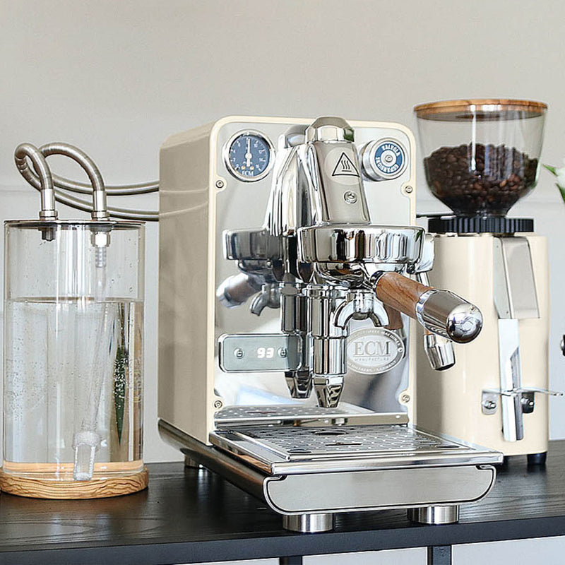 The ECM Puristika Semi Automatic Espresso Machine in a cream hue sits prominently on the counter, showcasing its sleek design. It features an E61 group head, chrome finish, pressure gauge, portafilter, and a transparent water reservoir. Accompanying it is a coffee grinder filled with beans, poised to perfect your brew.