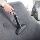 A person uses the Miele Wide Upholstery Nozzle Vacuum Cleaner Attachment SPD 20 to vacuum a gray upholstered chair. In the background, decorative vases with striped patterns can be seen.