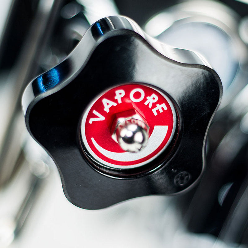 Close-up of a black, star-shaped valve handle featuring a red circular label at its center with the word "Vapore." A small metal knob extends from the middle of the label, and blurred metallic components of an ECM Mechanika VI Slim Espresso Machine - Demo, Unused can be seen in the background.