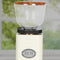 An ECM C-Manuale 54 Burr Grinder in cream is displayed indoors near a window, providing a blurred outdoor view. This on-demand coffee grinder features a wooden-topped bean hopper, and the silver logo plate complements the hopper's clear design, highlighting its modern aesthetic. Additionally, it offers precise graduate grind adjustments for perfect brews.