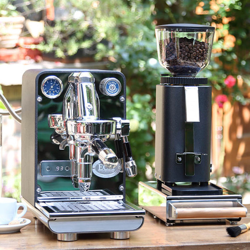 An ECM C-Manuale 54 Burr Grinder in Anthracite and a glossy espresso machine are positioned beside each other on a wooden table outdoors. Nearby, a white coffee cup complements the scene, with blurred greenery in the background setting up an ideal moment for your espresso enjoyment.