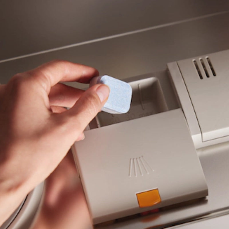 A hand places a Miele UltraTabs All in 1 Dishwasher Cleaning Tablet (Box of 60) into the detergent compartment of an open Miele dishwasher. The compartment lid, featuring a yellow release button and a faint Miele logo, is prominently shown. In the background, the dishwasher's interior glistens with promise.