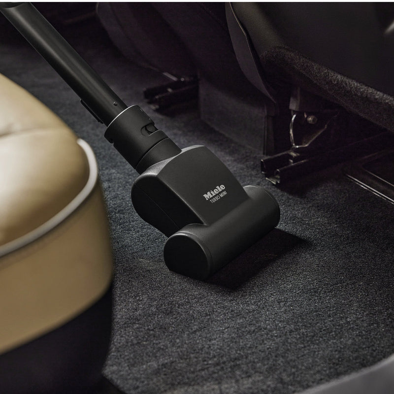 The Miele Handy TurboBrush Vacuum Cleaner Attachment STB 101 is expertly cleaning the black carpeted floor of a car interior, fitting perfectly between the front seat and passenger footwell. Its handle connects effortlessly to the eco-friendly Miele device's black nozzle, ensuring a comprehensive clean.