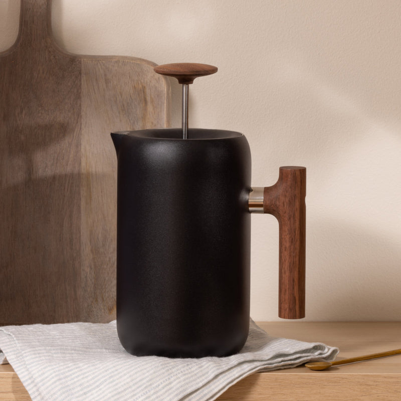 The Fellow Clara French Press (Matte Black + Walnut) by Fellow, which boasts barista-level functionality, features a wooden handle and plunger knob. It is placed on a light wooden surface with a gray striped cloth partially underneath it and a wooden cutting board in the background.