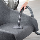 A person uses the Miele Wide Upholstery Nozzle Vacuum Cleaner Attachment SPD 20 to clean a gray upholstered chair with a Miele canister vacuum. The arm holding the eco-friendly vacuum is visible, and the surrounding area features a modern, minimalist design.